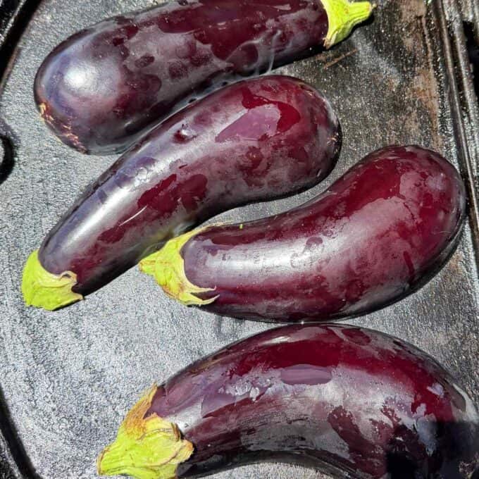 BBQ Miso Eggplant Four whole eggplants with shiny purple skin and green stems are placed on a dark surface.