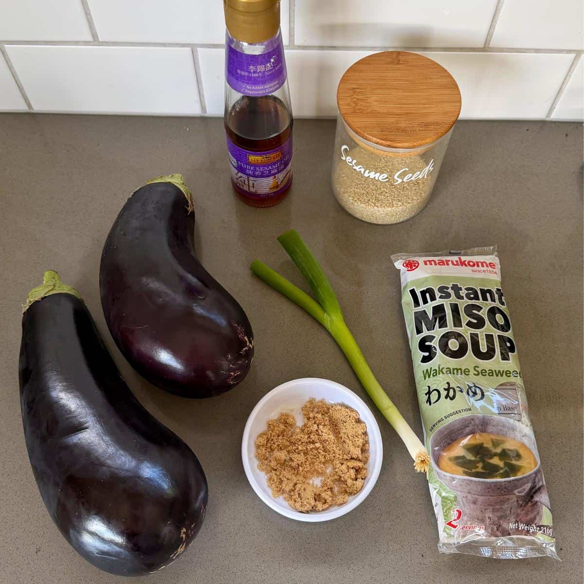 BBQ Miso Eggplant Two eggplants, green onions, brown sugar, instant miso soup, sesame seeds, and a bottle of sauce are arranged on a kitchen counter.