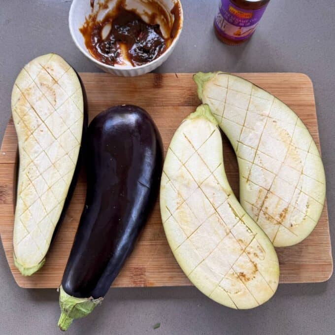 BBQ Miso Eggplant Three halved eggplants with crosshatch cuts and one whole eggplant on a wooden cutting board, with a bowl of sauce nearby.