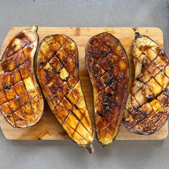 BBQ Miso Eggplant Four roasted eggplant halves with caramelized, crosshatched surfaces are arranged on a wooden cutting board.