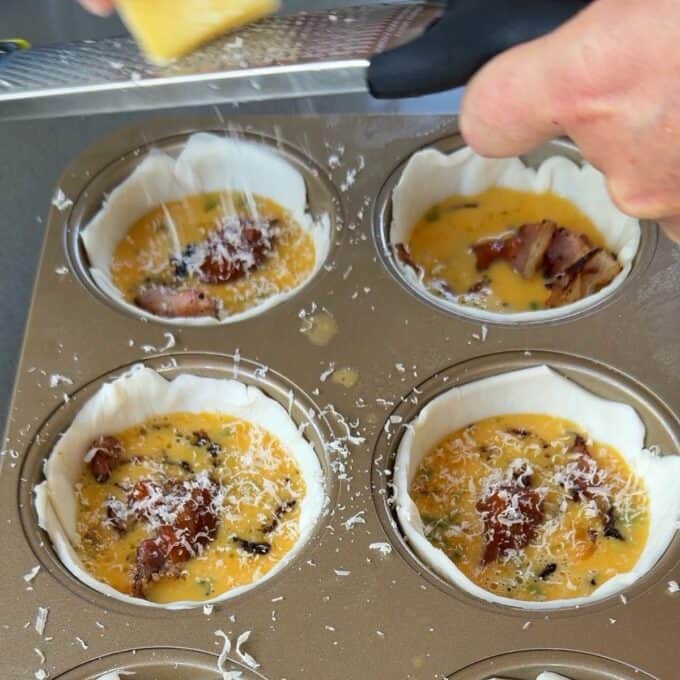 Bacon and Egg Picnic Pies Four unbaked mini quiches in a muffin tin with shredded cheese being grated on top of the egg, bacon, and vegetable filling.