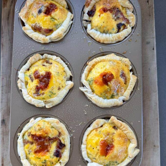 Bacon and Egg Picnic Pies Six baked mini quiches with golden, slightly browned tops sit in a metal muffin tray on a wooden surface.
