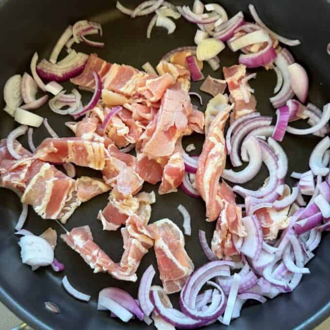 Bacon and Egg Picnic Pies Uncooked bacon strips and sliced red onions in a black frying pan.