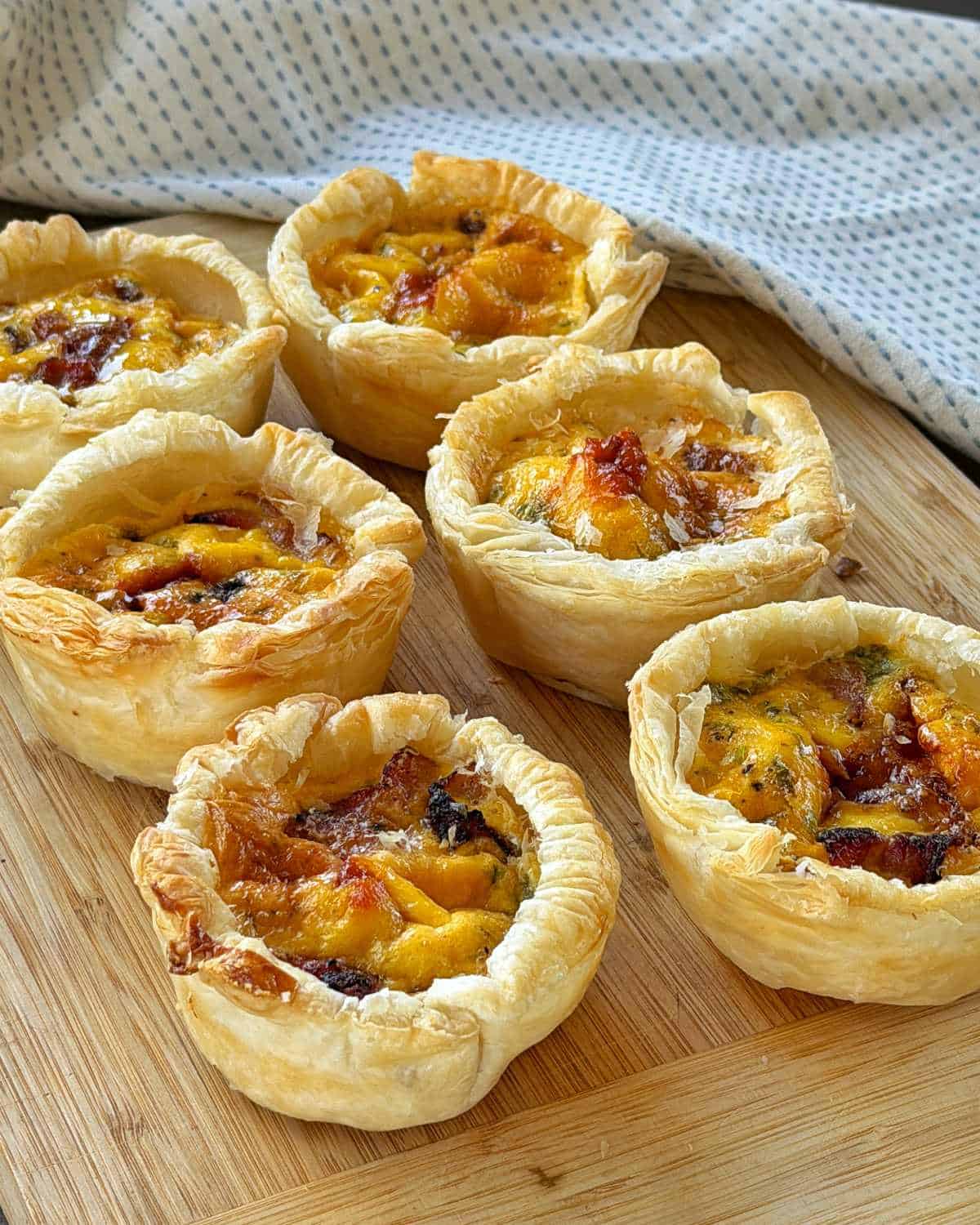 Bacon and Egg Picnic Pies Six individual mini quiches with golden, flaky pastry and visible cheese and vegetable fillings are arranged on a wooden cutting board with a cloth in the background.