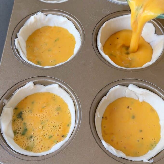 Bacon and Egg Picnic Pies Four muffin tin cups lined with pastry are being filled with an egg mixture, likely for making mini quiches or egg muffins.
