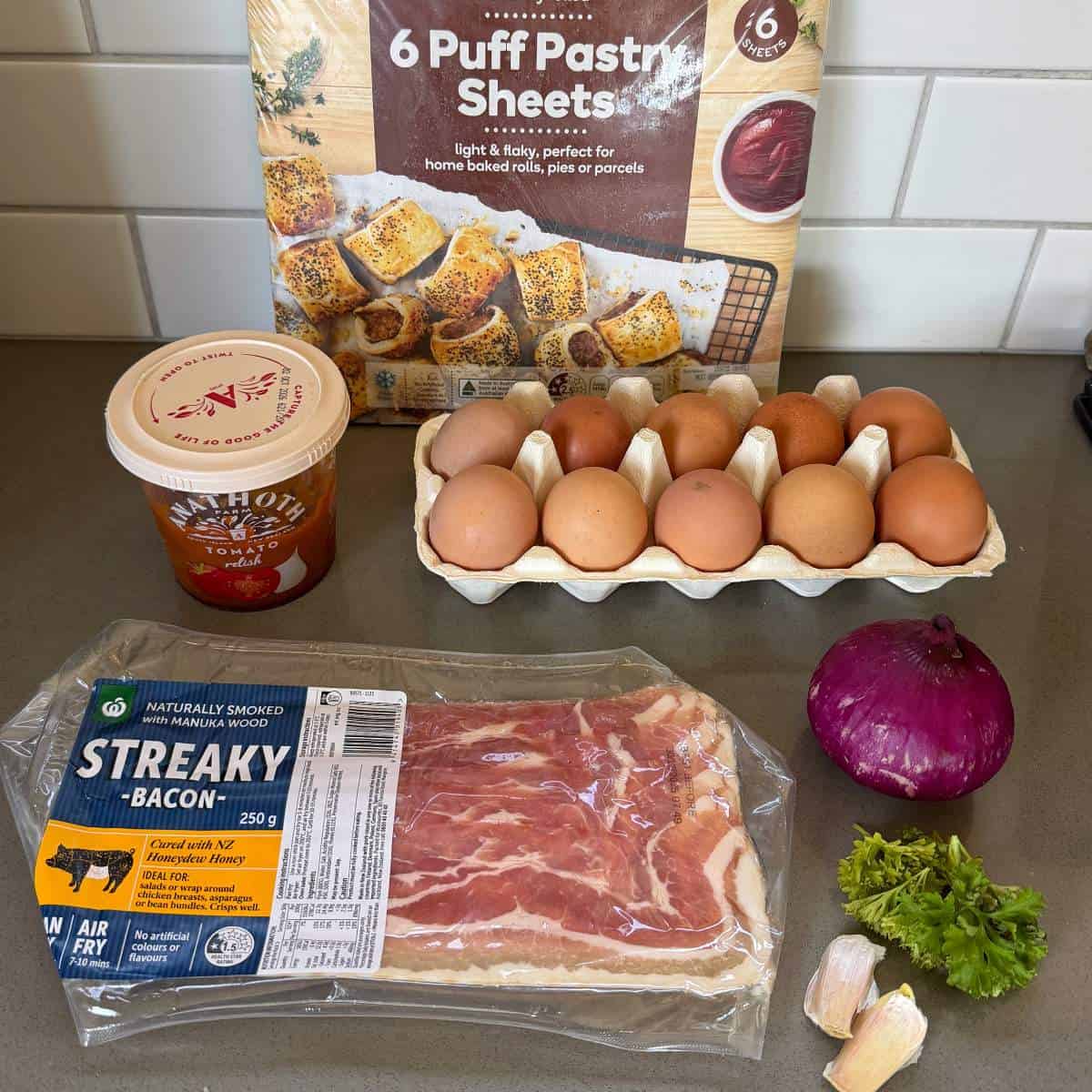 Bacon and Egg Picnic Pies A carton of eggs, a package of puff pastry sheets, a tub of double cream, a pack of streaky bacon, a red onion, two garlic cloves, and parsley are arranged on a kitchen counter.