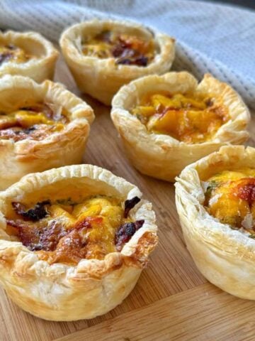 Six individual mini quiches with golden, flaky pastry and browned cheese on top, arranged on a wooden board with a cloth in the background.