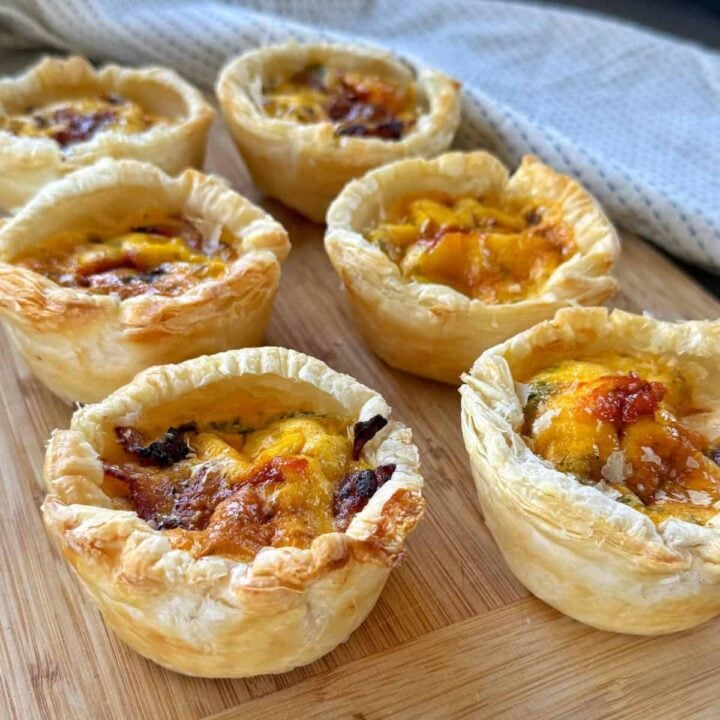 Six individual mini quiches with golden, flaky pastry and browned cheese on top, arranged on a wooden board with a cloth in the background.