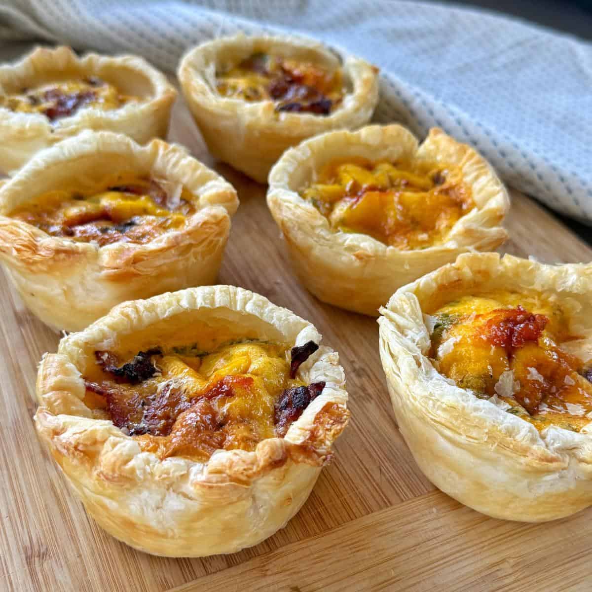 Bacon and Egg Picnic Pies Six individual mini quiches with golden, flaky pastry and browned cheese on top, arranged on a wooden board with a cloth in the background.