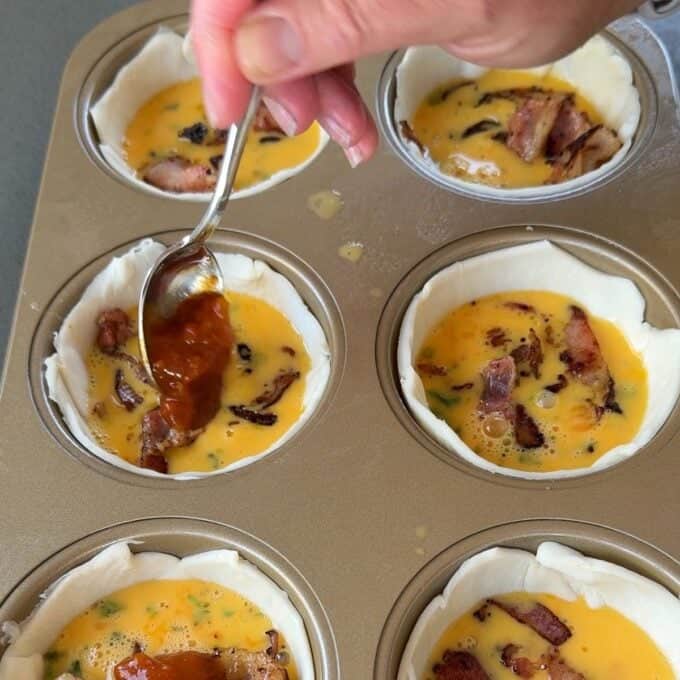 Bacon and Egg Picnic Pies A hand uses a spoon to add sauce to pastry-lined muffin tins filled with egg mixture and bacon pieces.