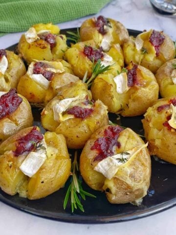 A black plate with smashed potatoes topped with cheese, herbs, and a dollop of cranberry sauce, garnished with fresh rosemary sprigs.
