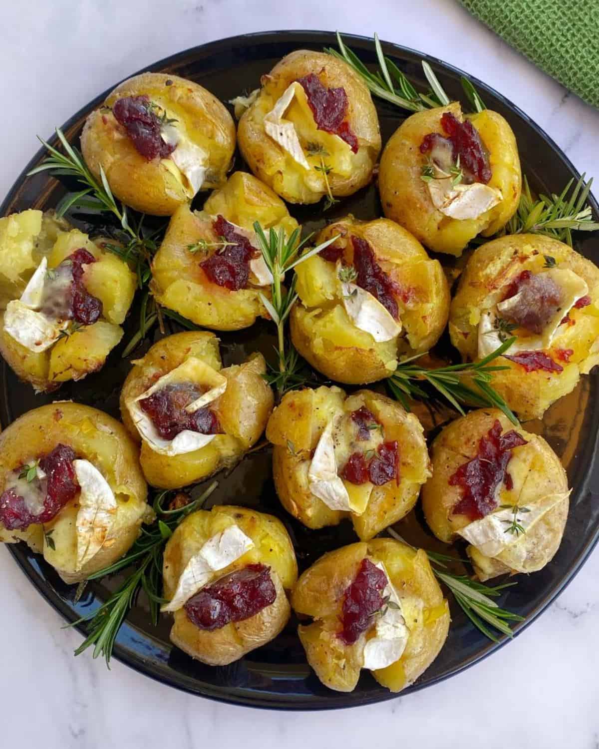 A black plate with smashed potatoes topped with cheese, sun-dried tomatoes, and herbs, garnished with fresh rosemary sprigs.