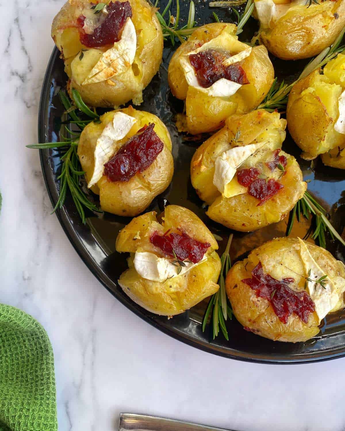 Brie and Cranberry Jacket Potatoes A black plate with smashed potatoes topped with melted cheese and cranberry sauce, garnished with rosemary sprigs on a marble surface.