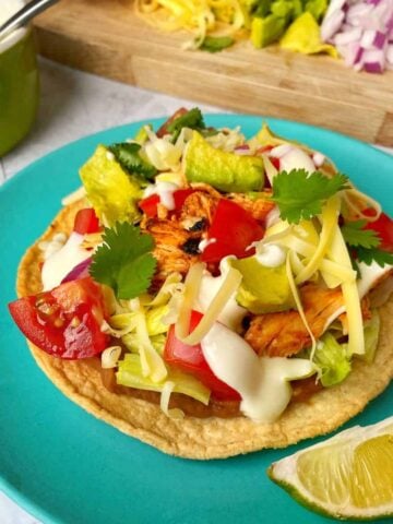 A chicken tostada topped with shredded cheese, tomatoes, avocado, cilantro, and sour cream on a blue plate with a lime wedge on the side.