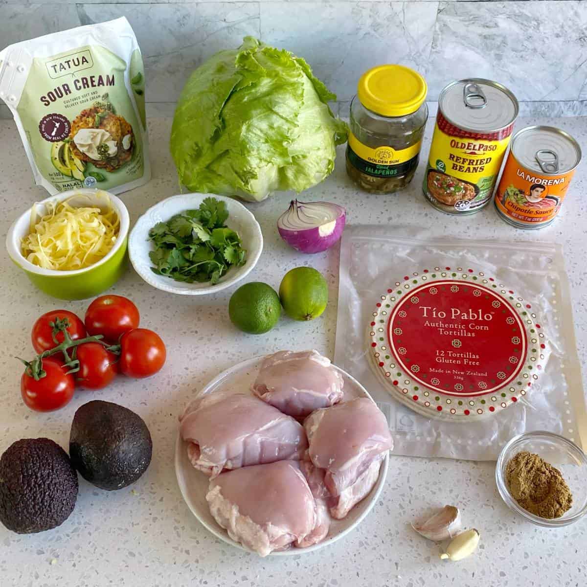 Chipotle Chicken Tostadas Various ingredients for making chicken tacos are arranged on a kitchen counter, including chicken thighs, lettuce, tortillas, avocado, canned beans, cheese, limes, tomatoes, and sour cream.