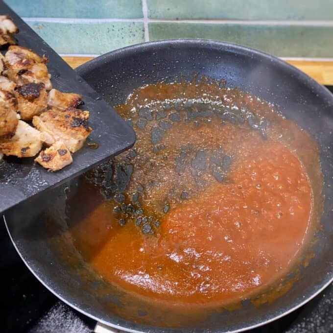 Chipotle Chicken Tostadas Cooked pieces of meat being added from a cutting board into a frying pan with simmering red sauce on a stove.