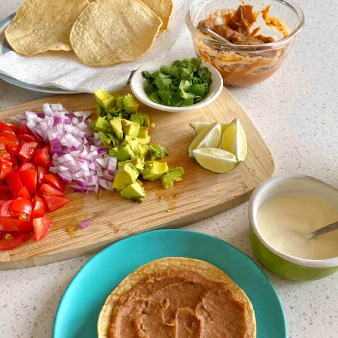 Chipotle Chicken Tostadas A kitchen counter with tostadas, refried beans, chopped tomatoes, red onion, avocado, cilantro, lime wedges, and a bowl of white sauce arranged for meal preparation.