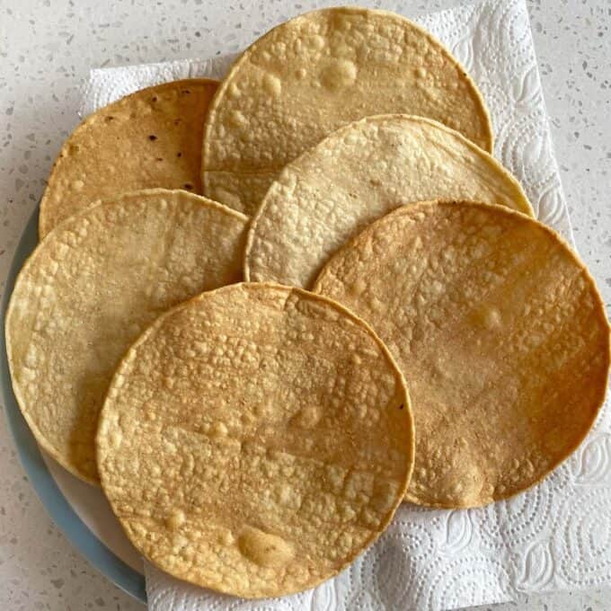 Chipotle Chicken Tostadas Six corn tortillas arranged on a white paper towel on a plate, set on a light-colored countertop.
