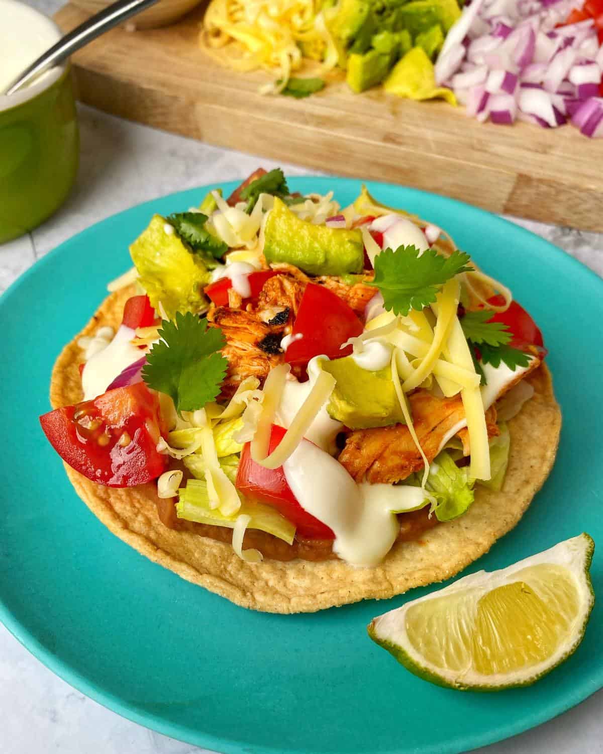 Chipotle Chicken Tostadas A tostada topped with grilled chicken, chopped tomatoes, lettuce, avocado, shredded cheese, cilantro, and sour cream, served on a blue plate with a lime wedge.