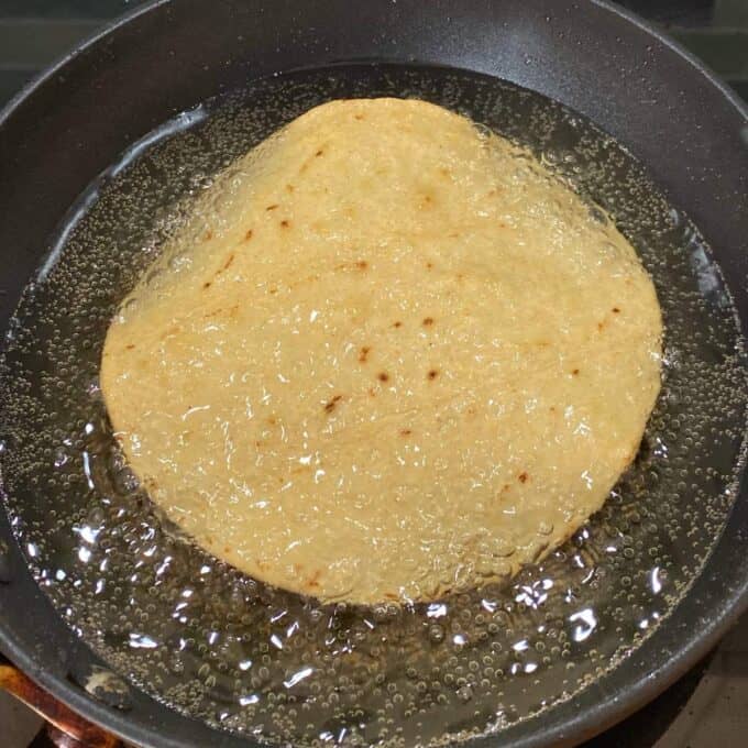 Chipotle Chicken Tostadas A round piece of dough is being deep-fried in hot oil in a black frying pan, with bubbles forming around it.