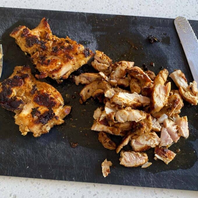 Chipotle Chicken Tostadas Two pieces of cooked, seasoned chicken—one whole and one sliced—on a black cutting board, with a fork and a knife nearby.