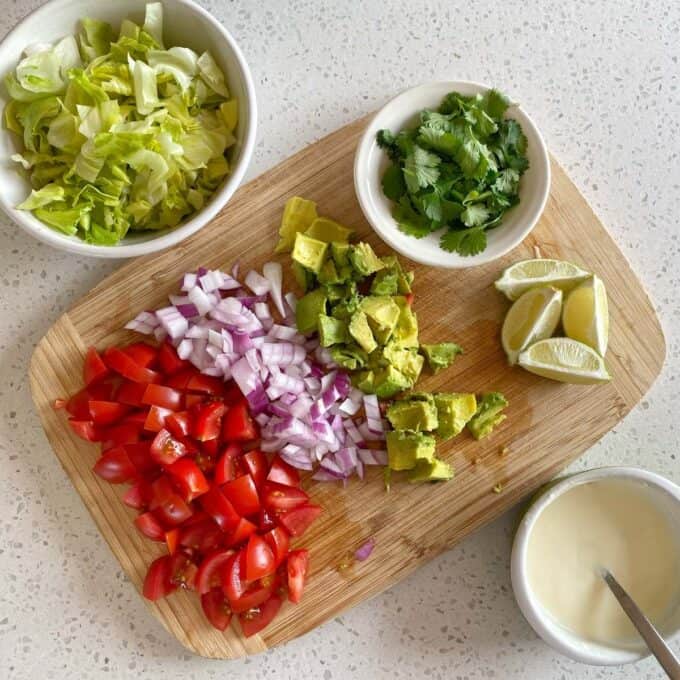 Chipotle Chicken Tostadas Chopped lettuce, tomatoes, red onion, avocado, lime wedges, cilantro, and a bowl of white sauce arranged on a cutting board and countertop.