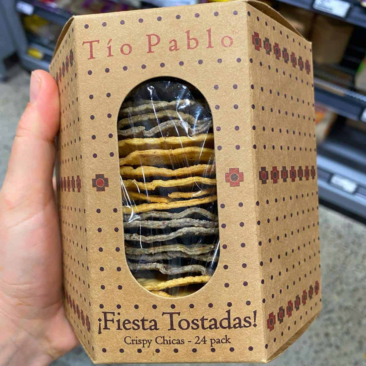 A hand holds a box of Tío Pablo Fiesta Tostadas, Crispy Chicas, 24-pack, with yellow and blue tostadas visible through a clear window.