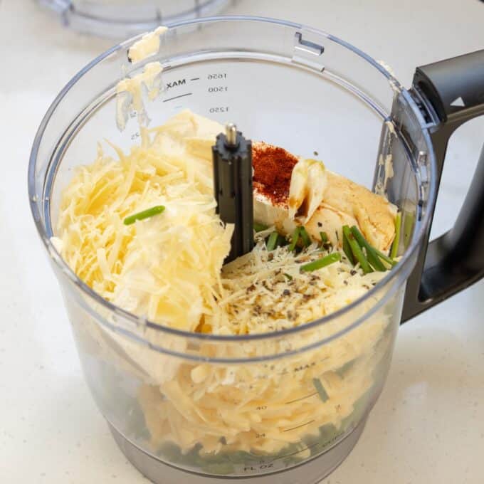 Christmas Cheeseball A food processor bowl filled with grated cheese, herbs, garlic, butter, and paprika on a white countertop, ready for mixing.