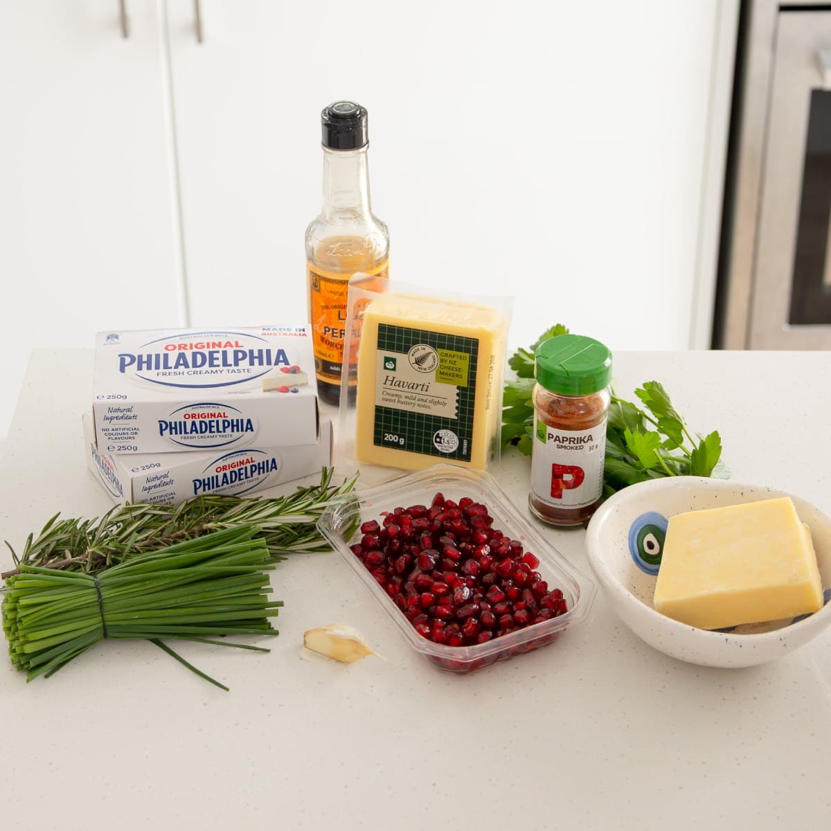 Christmas Cheeseball Ingredients on a white countertop include cream cheese, pomegranate seeds, fresh herbs, havarti cheese, butter, paprika, Worcestershire sauce, and parsley.