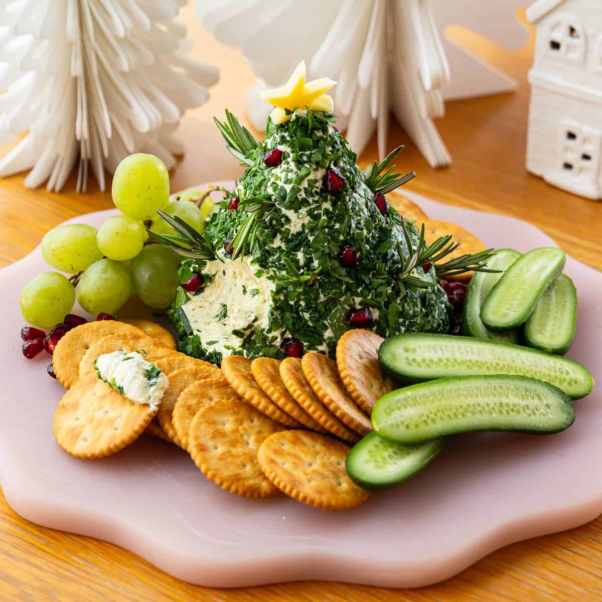 Christmas Cheeseball A cheese ball shaped like a Christmas tree, garnished with herbs and pomegranate seeds, is served with crackers, grapes, and sliced cucumbers on a pink plate.