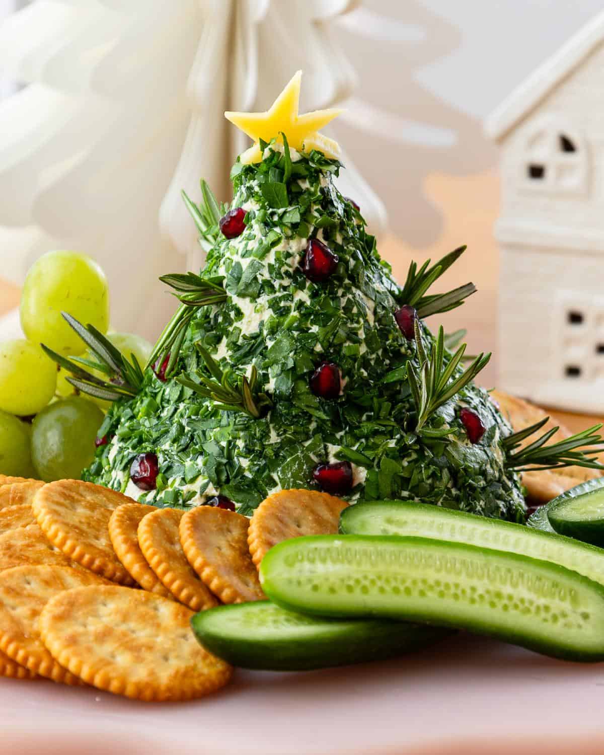 A Christmas tree-shaped cheese ball covered in herbs and garnished with rosemary, pomegranate seeds, a cheese star, served with crackers, cucumber slices, and green grapes.