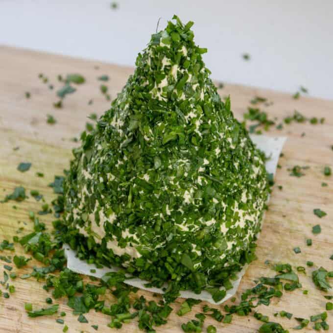 Christmas Cheeseball A pyramid-shaped mound of cheese coated with chopped fresh herbs sits on a wooden surface, with extra herbs scattered around.
