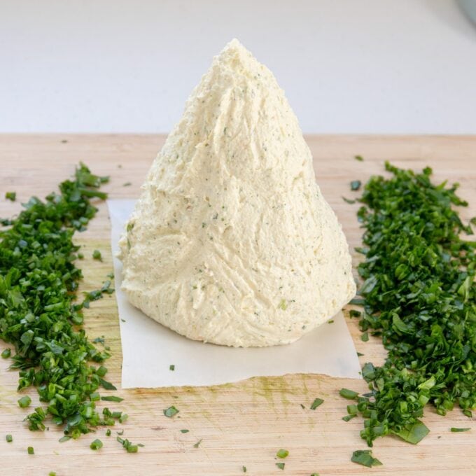 Christmas Cheeseball A cone-shaped mound of herbed cheese sits on parchment paper on a wooden board, with finely chopped herbs arranged on either side.