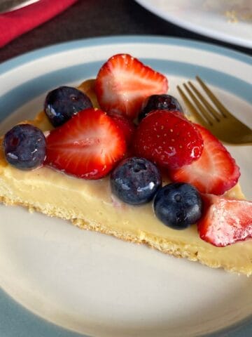 A slice of custard tart topped with fresh strawberries and blueberries on a plate with a gold fork.