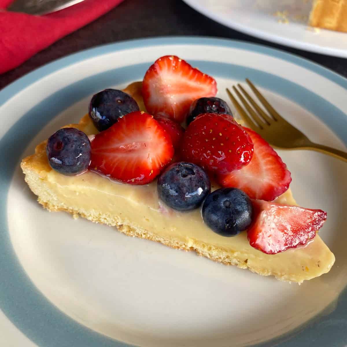 Fresh Berry Custard Tart A slice of custard tart topped with fresh strawberries and blueberries on a plate with a gold fork.