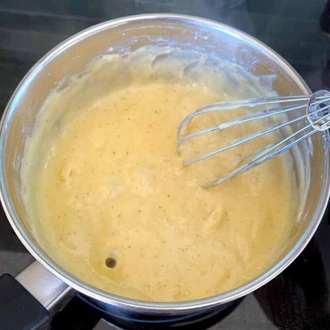 Fresh Berry Custard Tart A metal saucepan containing thick, creamy yellow sauce being whisked with a metal whisk on a stovetop.