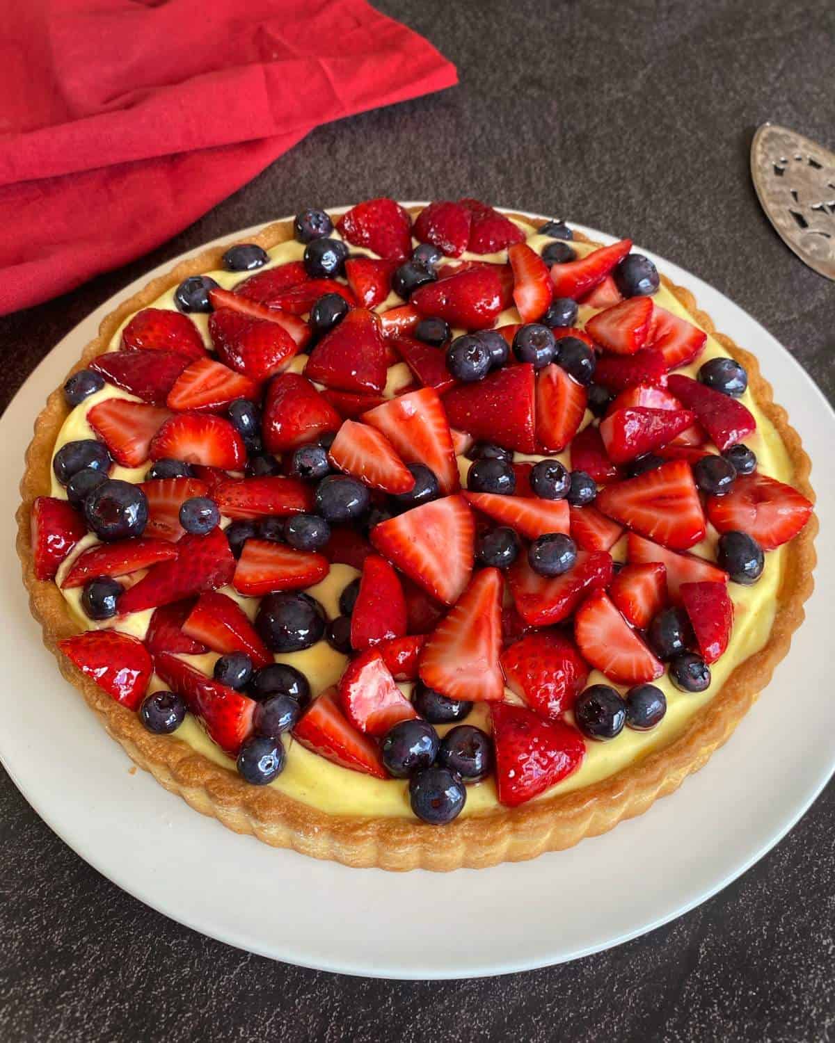 A fruit tart topped with sliced strawberries and whole blueberries sits on a white plate, with a red cloth nearby on a dark surface.