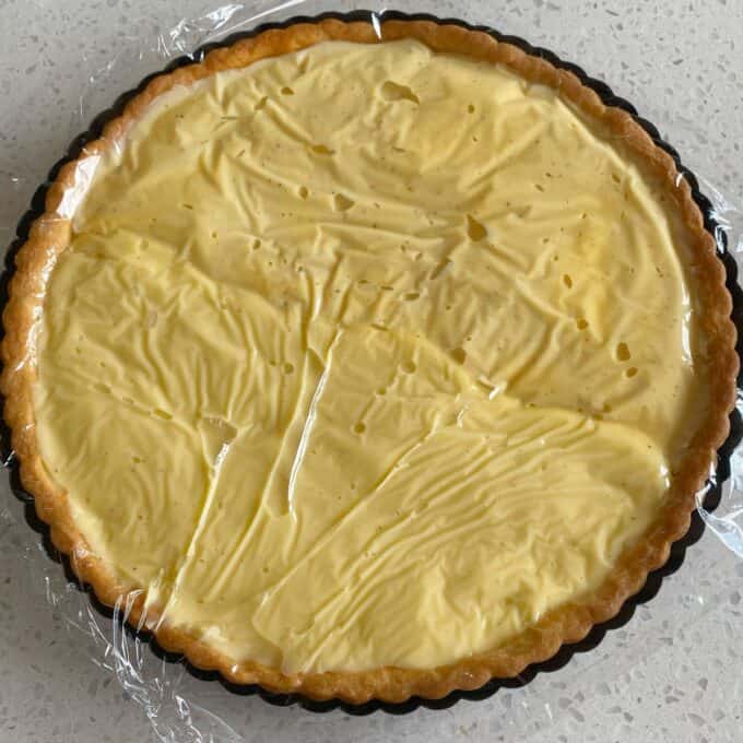 Fresh Berry Custard Tart A tart with a smooth, pale yellow custard filling in a pastry crust, covered loosely with clear plastic wrap, on a speckled countertop.