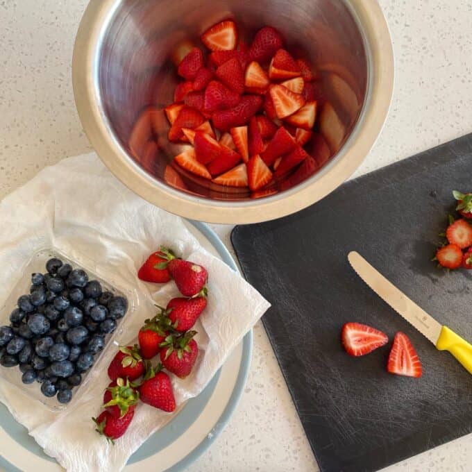 Fresh Berry Custard Tart A metal bowl of sliced strawberries, whole strawberries, blueberries on a plate, and a cutting board with a knife and strawberry tops on a light countertop.