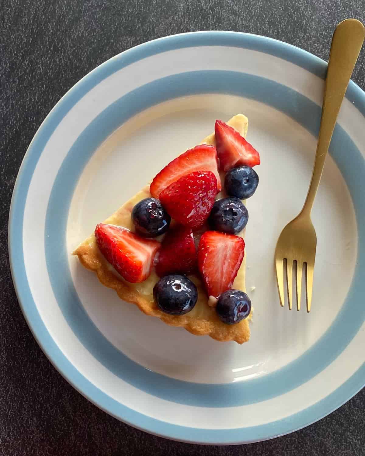 Fresh Berry Custard Tart A slice of fruit tart topped with strawberries and blueberries on a white plate with a blue rim, beside a gold fork.