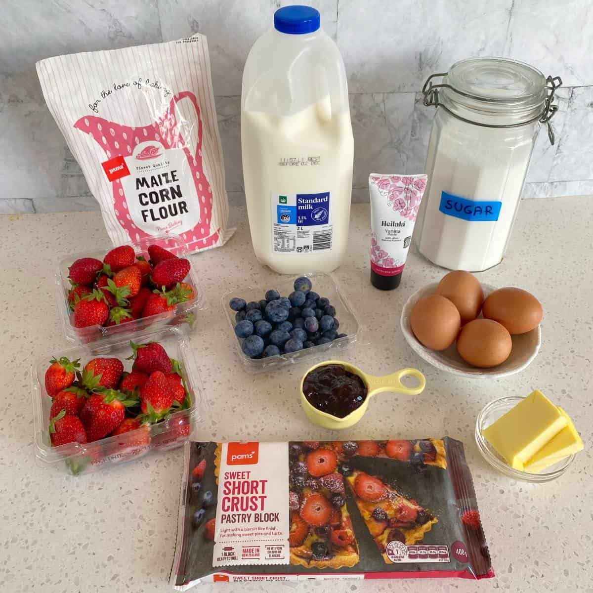 Fresh Berry Custard Tart A countertop displays corn flour, milk, sugar, eggs, butter, strawberries, blueberries, pastry block, jam, and cream in various containers.