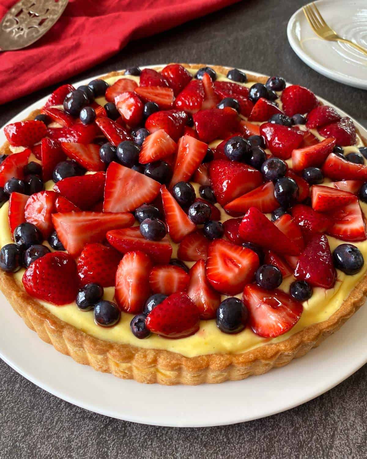 Fresh Berry Custard Tart A fruit tart topped with sliced strawberries and whole blueberries sits on a white plate, with a red cloth and plates in the background.