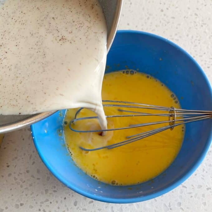 Fresh Berry Custard Tart Milk mixture is being poured from a metal bowl into a blue bowl with beaten eggs and a whisk on a light countertop.