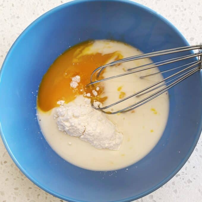 Fresh Berry Custard Tart A blue bowl containing an egg yolk, milk, and flour with a whisk resting inside, ready for mixing on a light countertop.