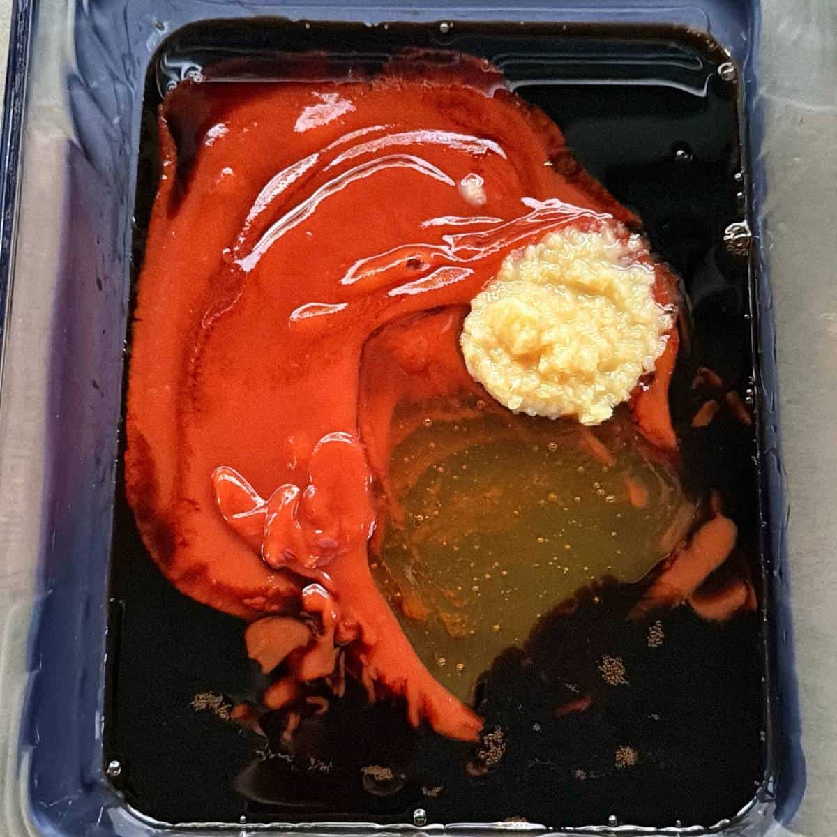 Honey Chicken Nibbles Rectangular container with a mixture of thick red sauce, dark brown liquid, and a mound of minced garlic or ginger.