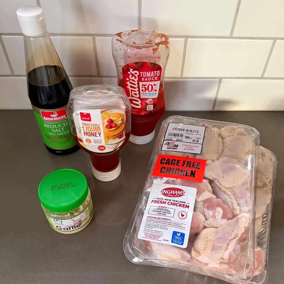 Honey Chicken Nibbles Five cooking ingredients on a kitchen counter: soy sauce, tomato sauce, liquid honey, minced garlic, and a pack of raw chicken wings. White tiled wall in the background.