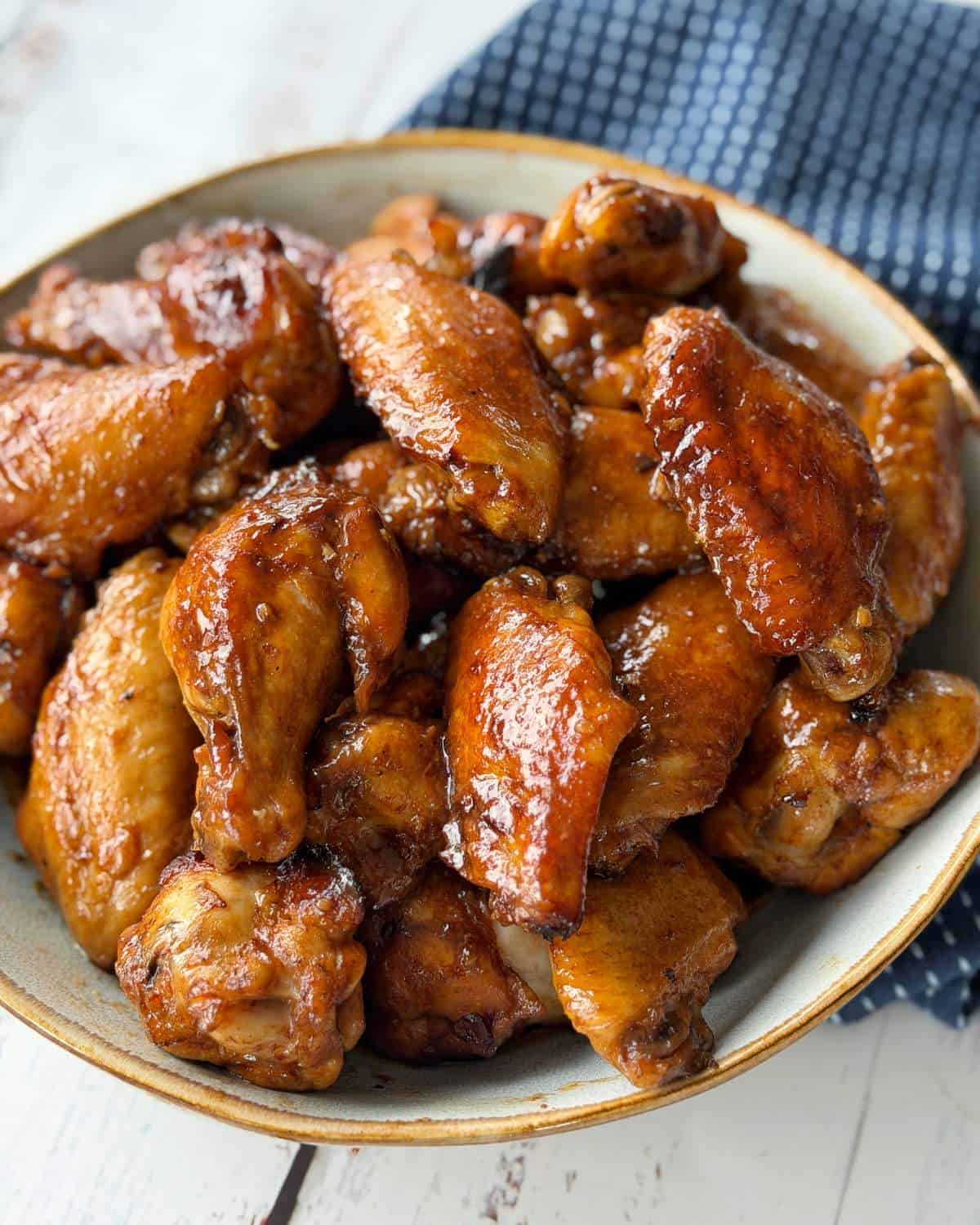A bowl filled with glazed, cooked chicken wings sits on a white surface next to a dark blue textured cloth.