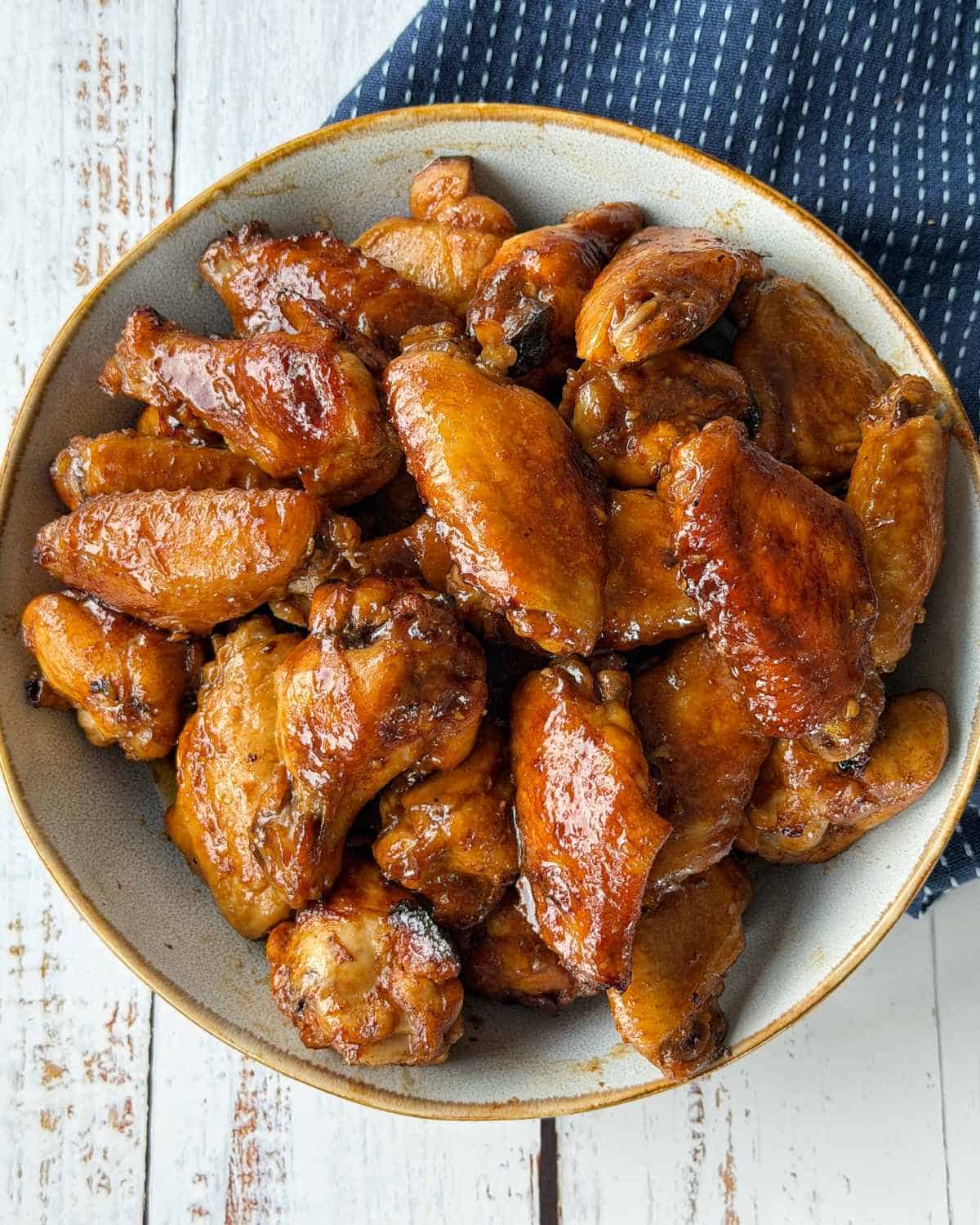 Honey Chicken Nibbles A bowl filled with glazed, cooked chicken wings sits on a white wooden surface next to a blue and white striped cloth.