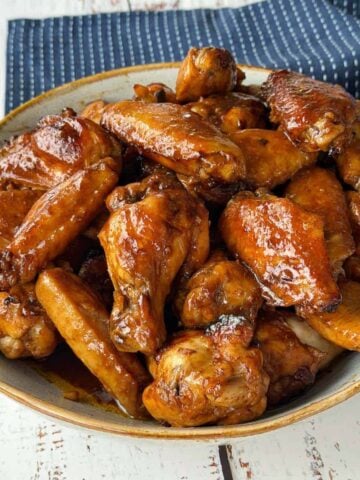 A bowl filled with glazed chicken wings sits on a white wooden table next to a dark blue cloth napkin with white dots.