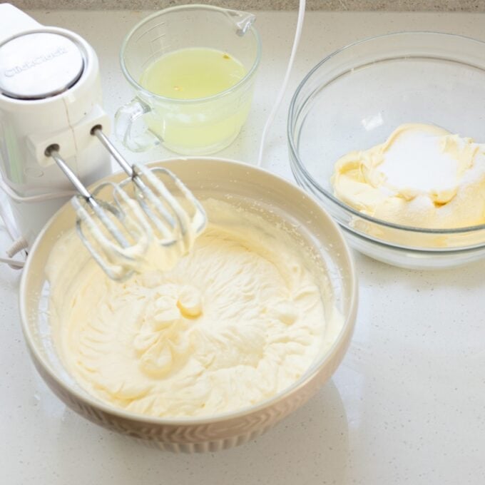 Limoncello Tiramisu A mixing bowl with creamed ingredients, an electric hand mixer, a bowl of sugar and butter, and a cup of liquid on a kitchen counter.